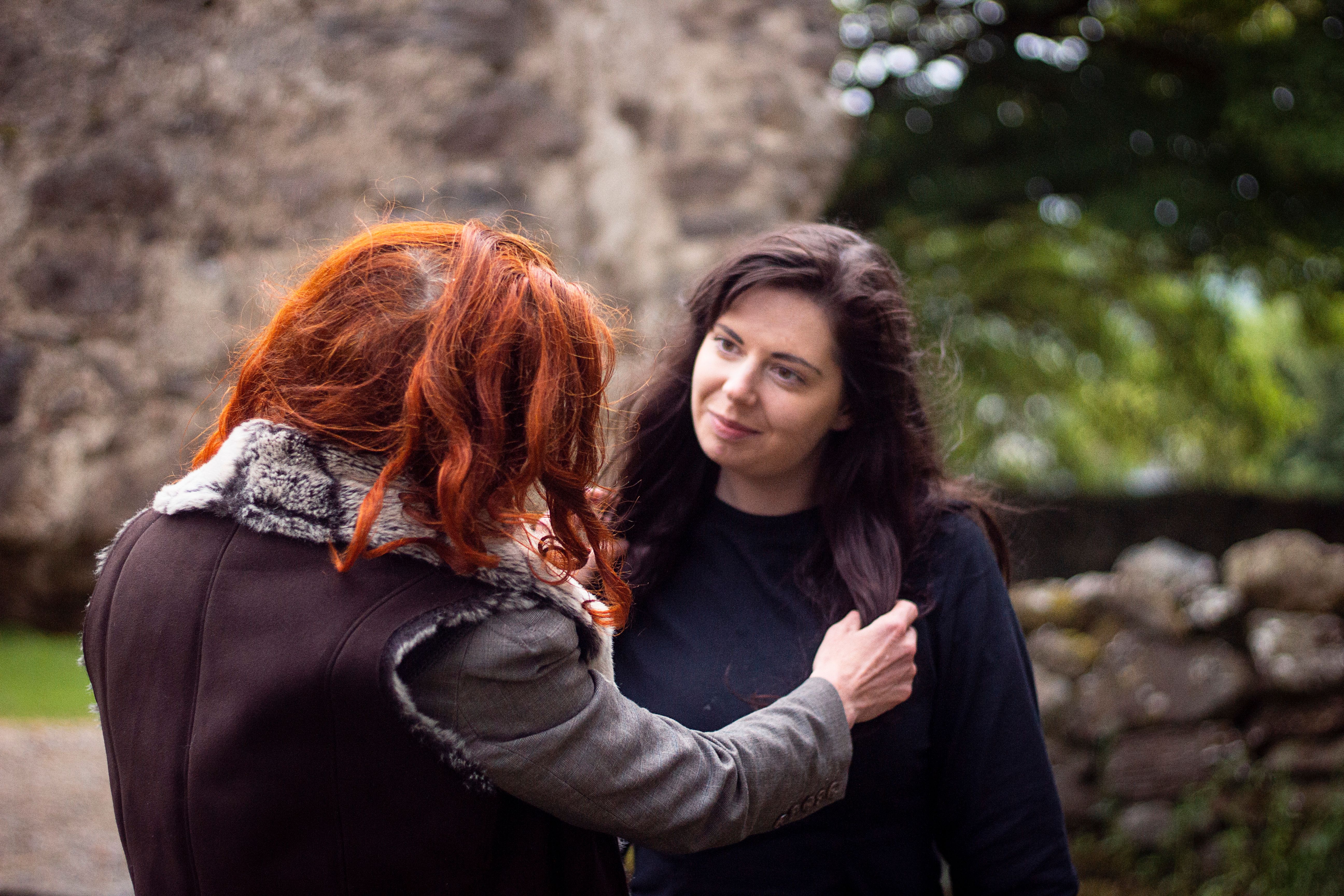 two female figures stand facing each other in front of a stone wall and natue backdrop